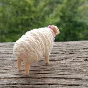 May include: A handcrafted sheep figurine with a fluffy, cream-colored wool body and a pink collar. The sheep has four wooden legs and is set against a blurred green background. The figurine is placed on a weathered wooden surface.