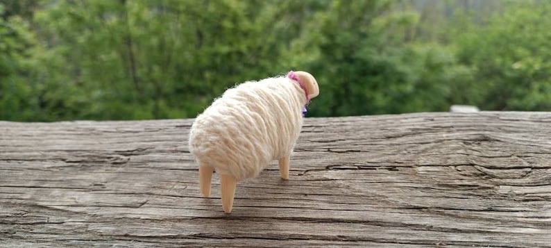 May include: A handcrafted sheep figurine with a fluffy, cream-colored wool body and a pink collar. The sheep has four wooden legs and is set against a blurred green background. The figurine is placed on a weathered wooden surface.