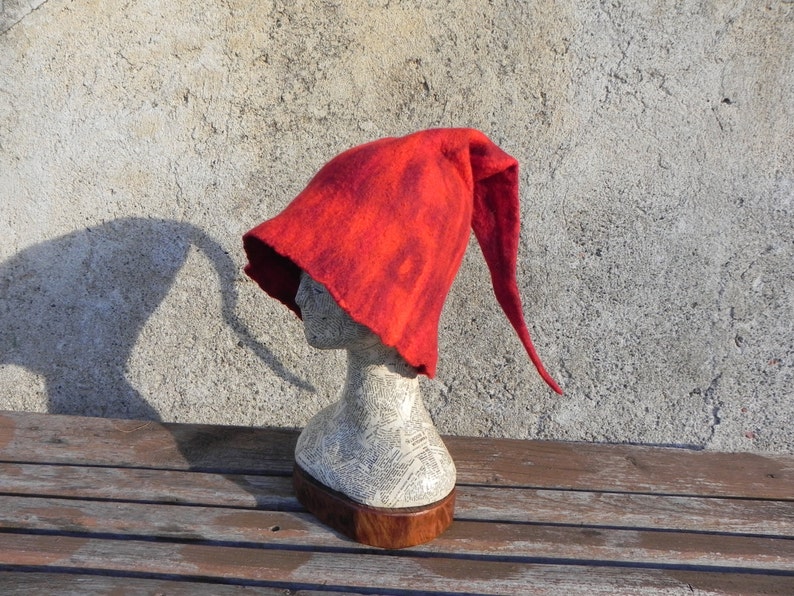 May include: A red felt hat with a long pointed crown. The hat is on a mannequin head and is sitting on a wooden surface.