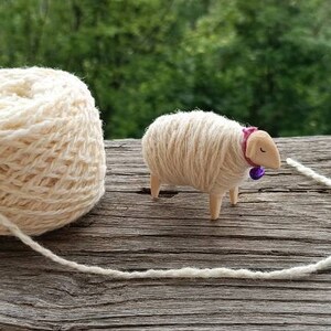 May include: A ball of cream-colored yarn next to a small wooden sheep figurine. The sheep has a pink collar with a purple bell. The yarn extends from the ball to the sheep, suggesting a crafting theme.