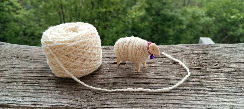 May include: A ball of cream-colored yarn next to a small wooden sheep figurine. The sheep has a pink collar with a purple bell. The yarn extends from the ball to the sheep, suggesting a crafting theme.