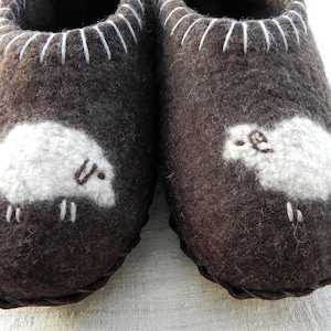 May include: A pair of brown wool slippers with white sheep appliques. The slippers have a white stitched trim around the top.