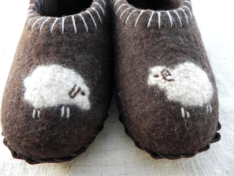 May include: A pair of brown wool slippers with white sheep appliques. The slippers have a white stitched trim around the top.