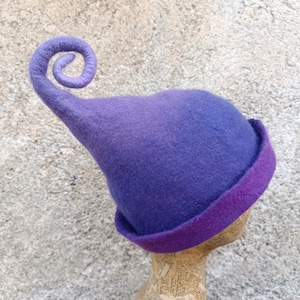 Può includere: Un cappello in feltro nelle tonalità del viola e del porpora, caratterizzato da una corona alta e appuntita che si arriccia a spirale sulla sommità. Il cappello presenta una tesa larga e ripiegata che sfuma dal viola chiaro alla base scura.