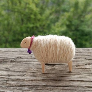 May include: A handcrafted wooden sheep figurine with a cream-colored wool body, light brown head and legs, and a purple bell collar. The sheep is standing on a weathered wooden surface, with a blurred green background.