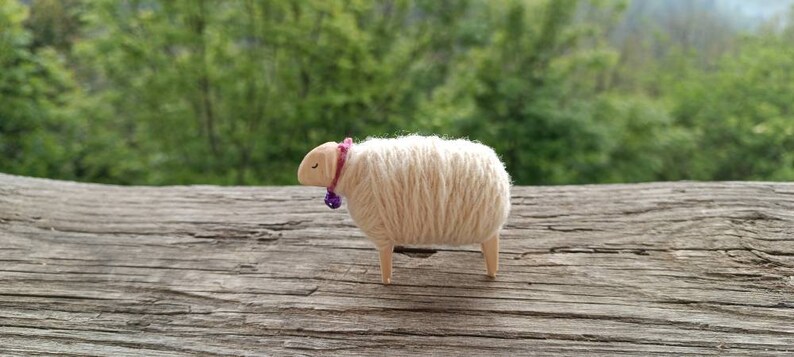 May include: A handcrafted wooden sheep figurine with a cream-colored wool body, light brown head and legs, and a purple bell collar. The sheep is standing on a weathered wooden surface, with a blurred green background.