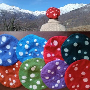 Può includere: Una collezione di baschi colorati con pois bianchi, a forma di funghi. I baschi sono in vari colori, tra cui rosso, blu, verde e viola. Una persona indossa un basco rosso con uno sfondo montuoso.