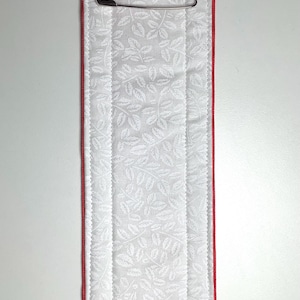May include: A rectangular, padded fabric item with a white leaf pattern, red trim, and blue and white star-patterned ends. The ends have metal clasps with the text "CAUTION MAGNETIC". A safety pin is attached.