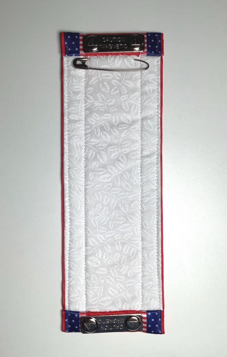 May include: A rectangular, padded fabric item with a white leaf pattern, red trim, and blue and white star-patterned ends. The ends have metal clasps with the text "CAUTION MAGNETIC". A safety pin is attached.