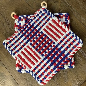 May include: Two star-shaped woven pot holders in red, white, and blue. Each pot holder has a wooden ring for hanging. The woven design creates a textured surface. The pot holders are on a wooden surface.