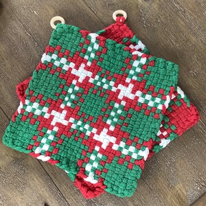 May include: Two woven pot holders in green, red, and white with wooden hanging loops. The pot holders feature a plaid pattern. The colors are arranged in a woven design, creating a festive and decorative look. The pot holders are square-shaped.