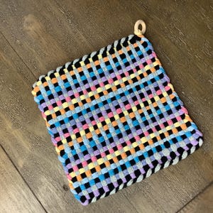 May include: A colorful woven pot holder with a black border and a wooden ring attached. The pot holder is made of a variety of colors, including pink, blue, yellow, and orange.