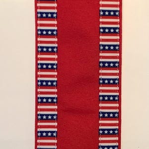May include: A red ribbon with a central panel and borders featuring a repeating pattern of white stripes, blue stars, and red stripes. The ribbon is likely used for decorative purposes or crafts.