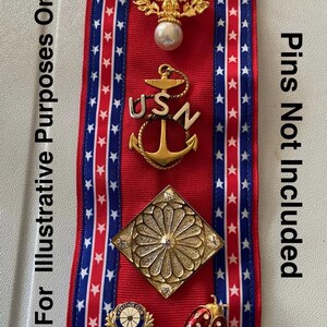 May include: A red ribbon with blue and white star-patterned stripes, adorned with various pins. Pins include a gold eagle with a pearl, a USN anchor, a floral design, a ladybug, and other emblems. Text on the side reads "For Illustrative Purposes Only" and "Pins Not Included."