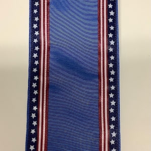 May include: Blue ribbon with a central blue stripe and borders featuring white stars on a navy blue background, with red and white stripes. The ribbon is likely for decoration or awards.