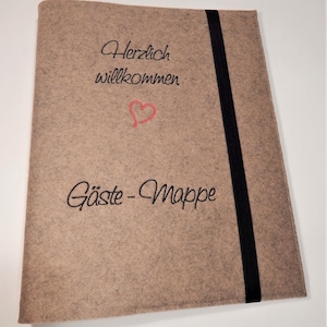 Könnte beinhalten: Hellbraune Gästemappe aus Filz mit schwarzem Gummibandverschluss. Der Umschlag zeigt einen schwarzen Schriftzug "Herzlich willkommen" über einem roten Herz und "Gäste-Mappe".