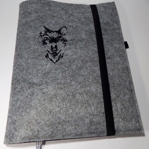 Könnte beinhalten: Eine graue Filz-Notizbuchhülle mit einem schwarzen Gummibandverschluss. Die Hülle hat ein gesticktes Wolfskopf-Design. Ein kleines kariertes Band ist am unteren Rand befestigt.