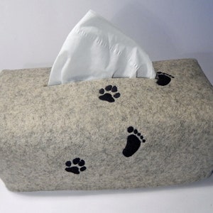 Könnte beinhalten: Ein rechteckiger, hellgrauer Taschentuchbox-Bezug aus Filz. Schwarze Pfoten- und Fußabdrücke sind auf die Vorderseite gestickt. Weiße Taschentücher sind sichtbar und kommen aus der oberen Öffnung heraus. Ein dekorativer Artikel für Tierliebhaber.