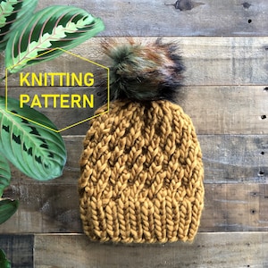 Puede incluir: Un gorro de punto amarillo mostaza con un gran pompón esponjoso hecho de piel sintética negra, marrón y verde. El gorro está hecho con un hilo grueso y tiene un patrón de punto texturizado. El gorro está fotografiado sobre un fondo de madera rústico.