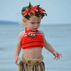 May include: A child's handmade Hawaiian costume. The outfit includes a red and white striped crochet top, a tan and brown fringe skirt, and a floral headpiece. The child is standing on a sandy beach.
