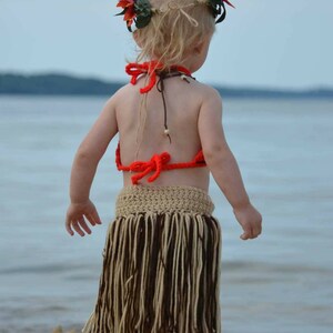 May include: A child wearing a handmade Hawaiian-themed outfit stands on a sandy beach. The outfit includes a crochet top, a fringed skirt, and a floral headpiece. The skirt is beige and brown, and the top is bright orange.