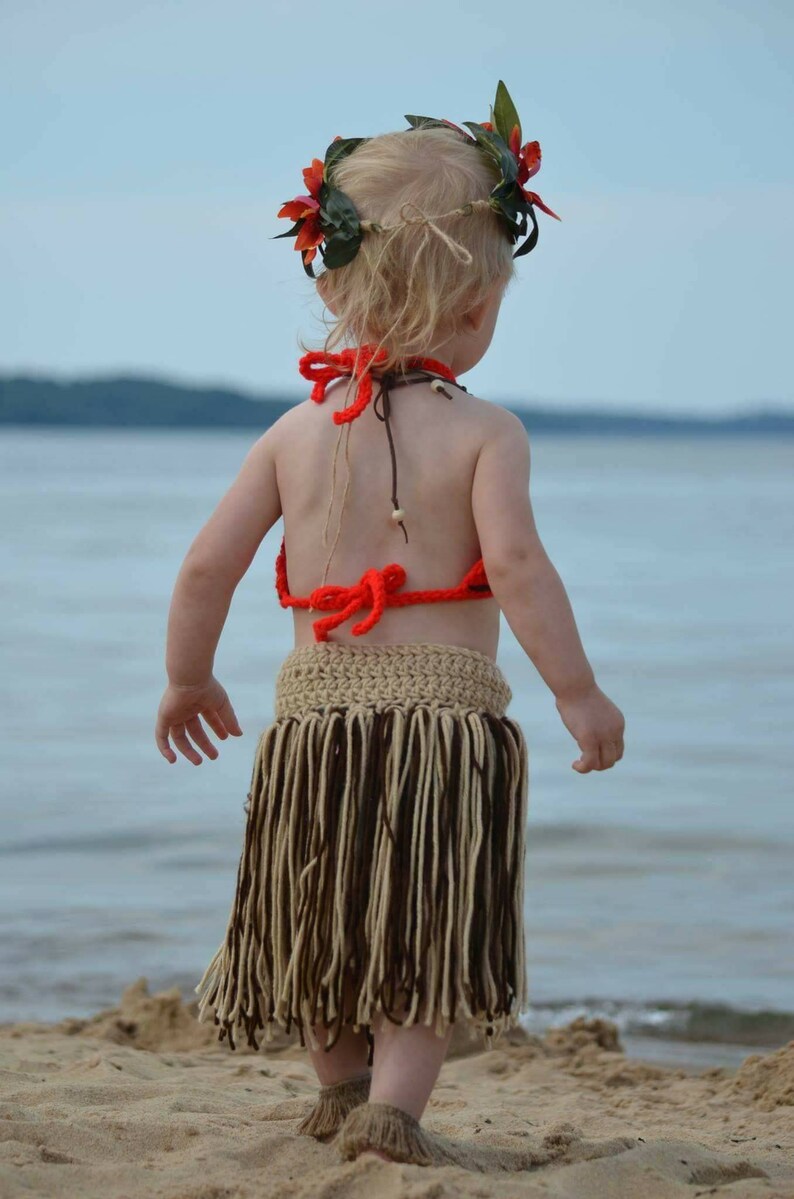 May include: A child wearing a handmade Hawaiian-themed outfit stands on a sandy beach. The outfit includes a crochet top, a fringed skirt, and a floral headpiece. The skirt is beige and brown, and the top is bright orange.
