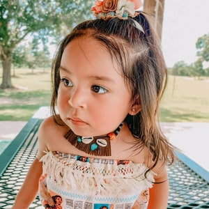 May include: A young child wearing a colorful outfit with a floral hair accessory and a necklace. The outfit features a print of a cartoon character. The necklace includes shells and turquoise beads. The child is sitting outdoors.