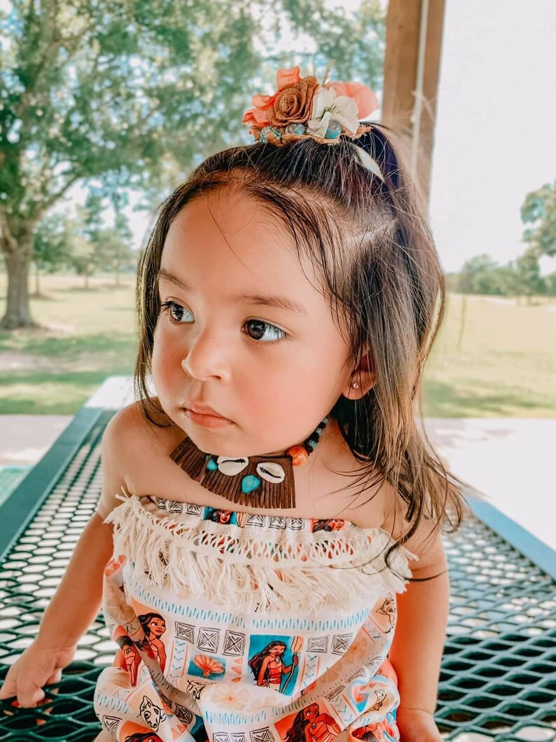May include: A young child wearing a colorful outfit with a floral hair accessory and a necklace. The outfit features a print of a cartoon character. The necklace includes shells and turquoise beads. The child is sitting outdoors.
