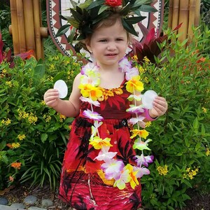 May include: A young child wearing a red tropical print dress, a flower lei, and a leafy headpiece. The child is holding two white circular objects and wearing silver sandals. A sign in the background reads "Spirit of Aloha Dinner Show."