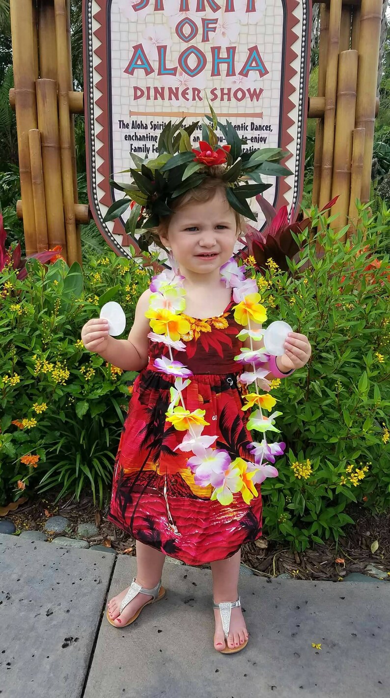 May include: A young child wearing a red tropical print dress, a flower lei, and a leafy headpiece. The child is holding two white circular objects and wearing silver sandals. A sign in the background reads "Spirit of Aloha Dinner Show."