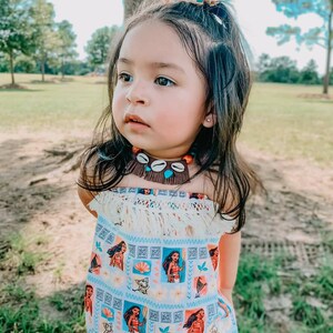May include: A young child wearing a strapless romper with a light blue background and a repeating pattern of a cartoon character. The romper has a fringe trim and is paired with a shell necklace and a floral hair accessory.