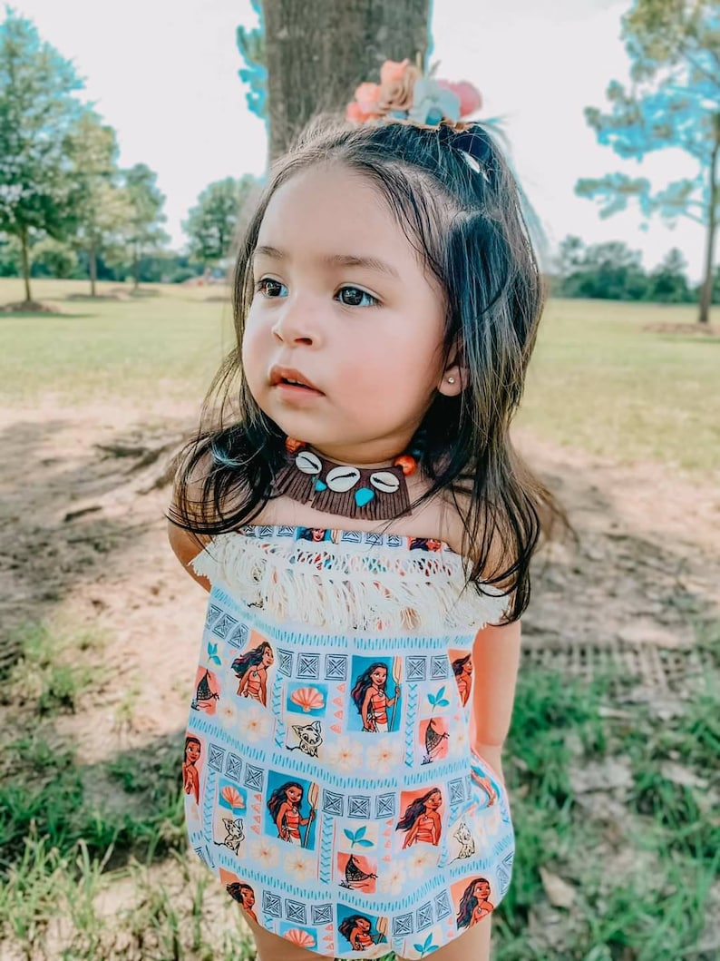 May include: A young child wearing a strapless romper with a light blue background and a repeating pattern of a cartoon character. The romper has a fringe trim and is paired with a shell necklace and a floral hair accessory.