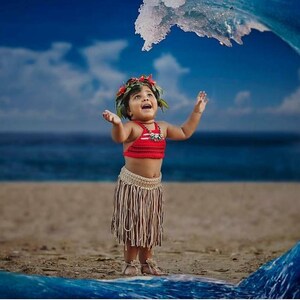 May include: A child in a red top and fringed skirt stands on a sandy beach with arms raised. A floral headpiece and necklace complete the outfit. A large wave arches over the child, with the ocean and sky in the background.