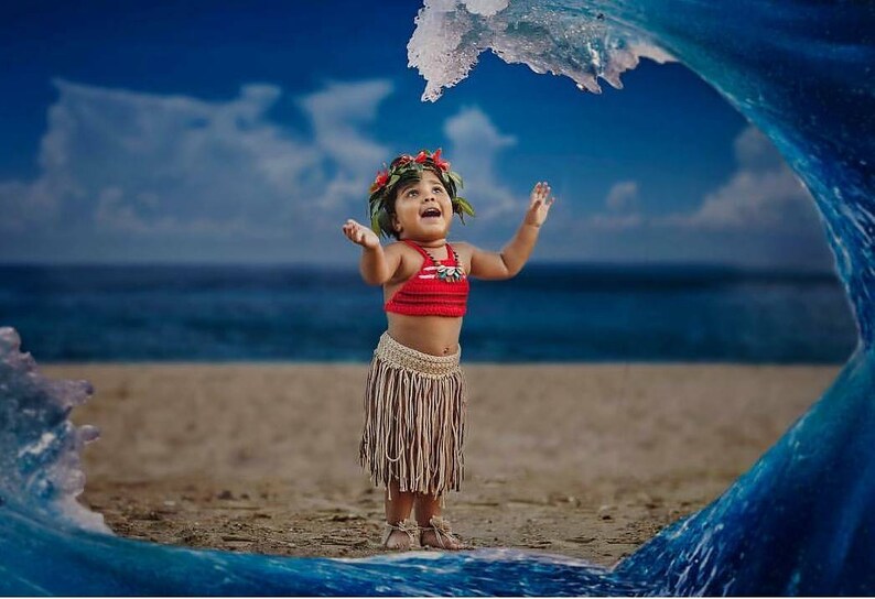 May include: A child in a red top and fringed skirt stands on a sandy beach with arms raised. A floral headpiece and necklace complete the outfit. A large wave arches over the child, with the ocean and sky in the background.