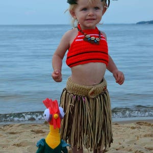 May include: A young child in a red and black crochet top, a tan skirt, and a floral headpiece stands on a sandy beach. A plush chicken toy is at their feet. The child is wearing a shell necklace.