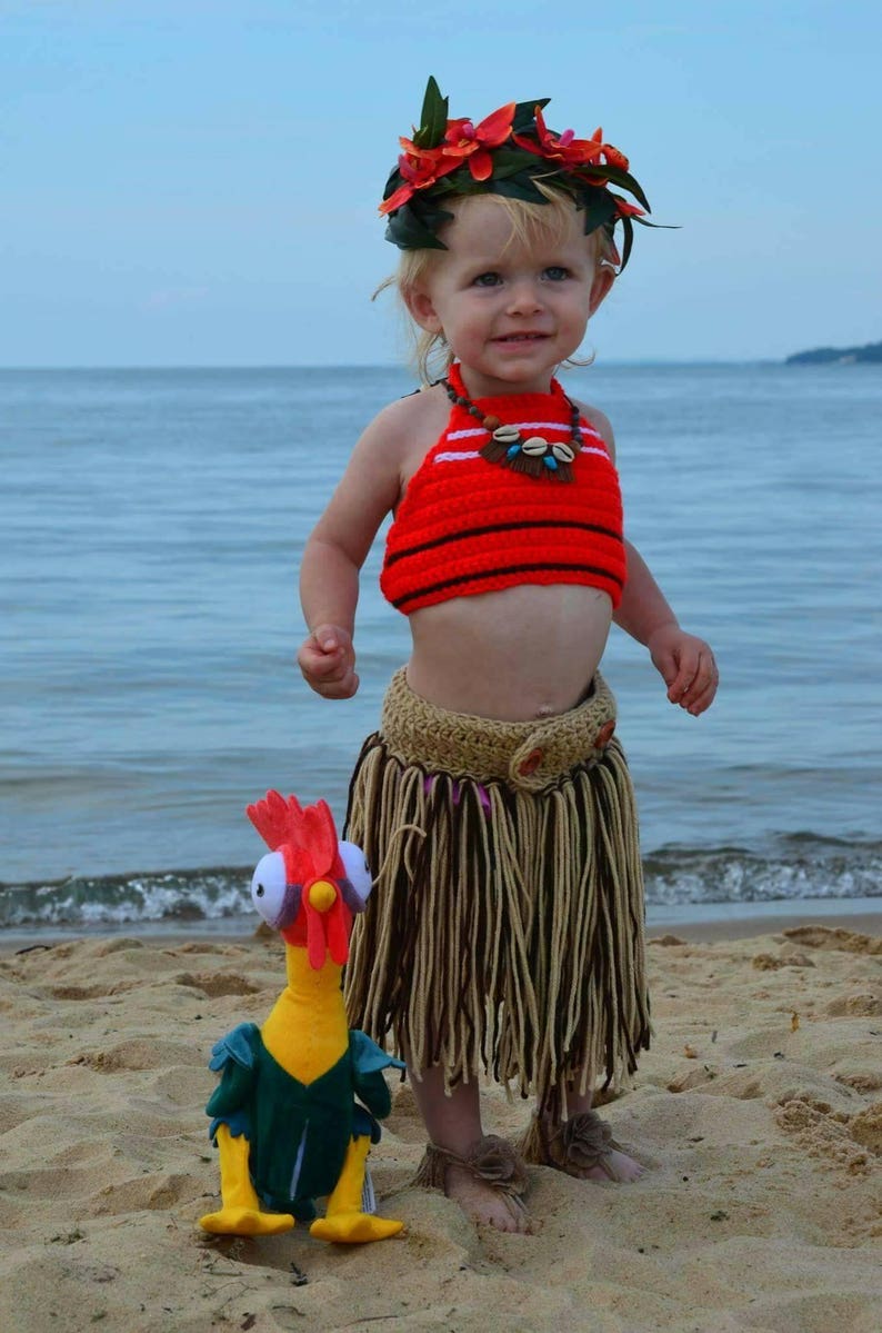 May include: A young child in a red and black crochet top, a tan skirt, and a floral headpiece stands on a sandy beach. A plush chicken toy is at their feet. The child is wearing a shell necklace.