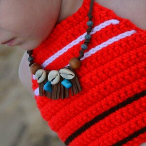 May include: A handmade red crochet halter top with white stripes and brown accents. The top features a necklace with seashells, wooden beads, and turquoise stones. The halter top is a unique, handcrafted piece.