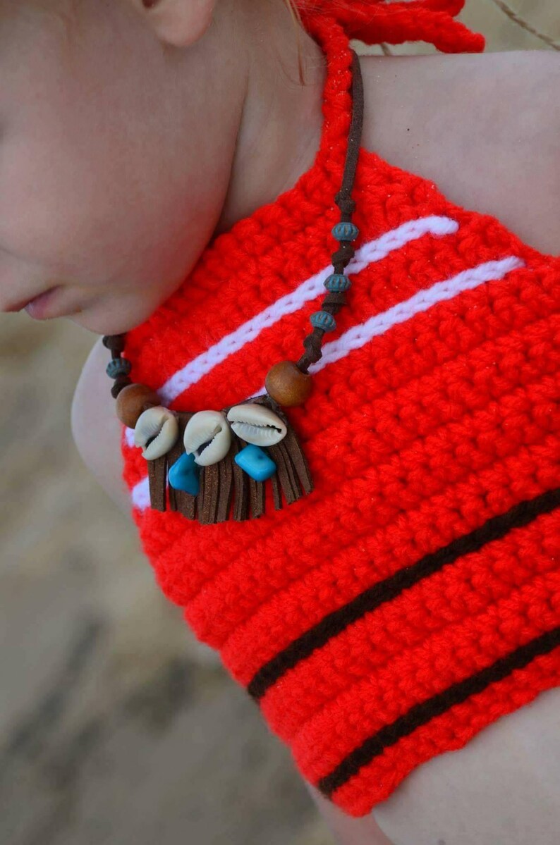 May include: A handmade red crochet halter top with white stripes and brown accents. The top features a necklace with seashells, wooden beads, and turquoise stones. The halter top is a unique, handcrafted piece.
