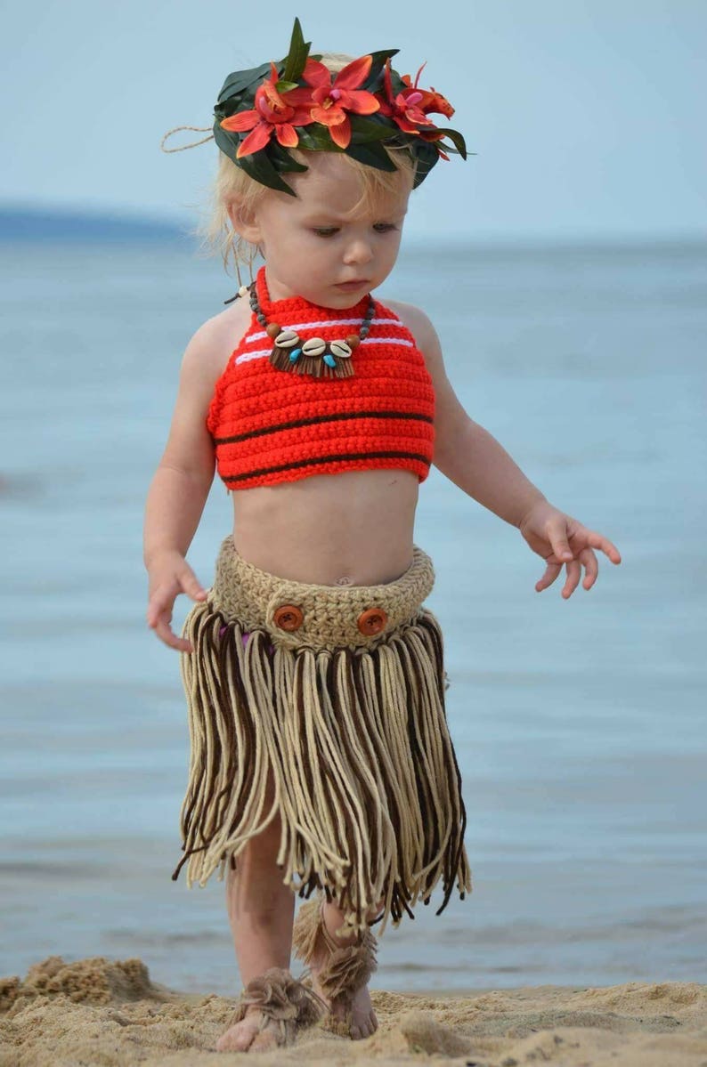 May include: A child's handmade Hawaiian costume. The outfit includes a red and white striped crochet top, a tan and brown fringe skirt, and a floral headpiece. The child is standing on a sandy beach.