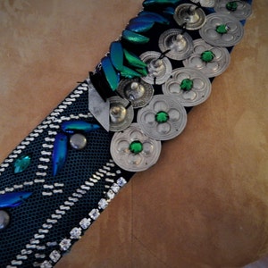 Puede incluir: Un cinturón azul oscuro y plateado con gemas verdes y alas de escarabajo iridiscentes. El cinturón está hecho de una tela tejida y tiene un diseño decorativo.