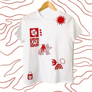 May include: White t-shirt with red graphic designs. The shirt features a sun, flower, geometric shapes, a dog-like creature, a bird, a rainbow, and a house. The shirt is hanging on a wooden hanger.