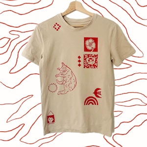 May include: A beige t-shirt featuring red graphic designs, including a cat, floral motifs, geometric shapes, and a house. The tee is displayed on a wooden hanger against a white backdrop with red line art.
