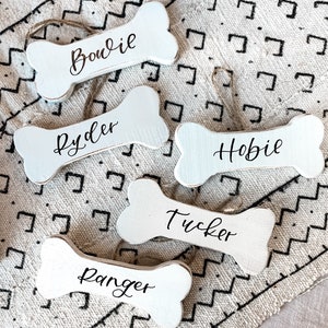 May include: Five white wooden dog bone ornaments with black lettering. The names on the ornaments are Bowie, Ryder, Hobie, Tucker, and Ranger.