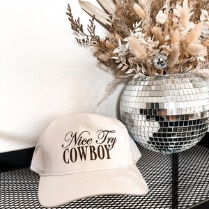 May include: White trucker hat with brown mesh back and the text "Nice Try COWBOY" in a cursive font.