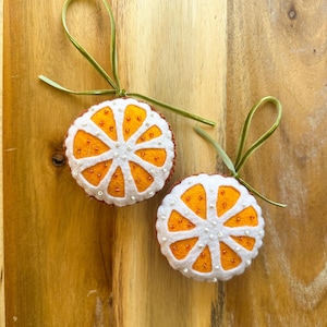 May include: Two felt orange slice ornaments with white segments, orange centers, and small bead accents. Each ornament has a green ribbon loop for hanging. The ornaments are displayed on a wooden surface.