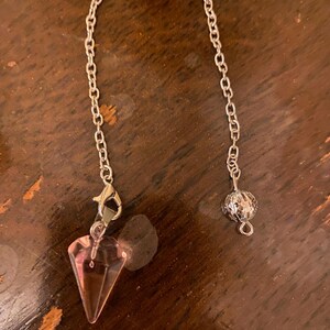 May include: A pendulum featuring a silver chain and a pink, faceted crystal point. The chain is connected to a small, decorative silver ball. This pendulum is designed for divination and spiritual use.
