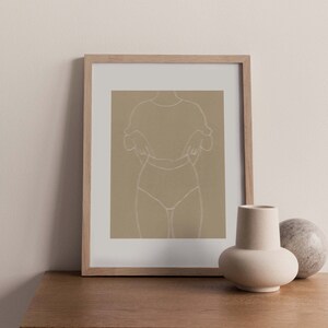 May include: A framed print of a line drawing of a person in a simple outfit, with a light brown background. The print is on a wooden shelf with a white vase and a round stone object.