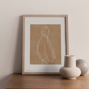 Peut inclure: Une impression encadr&eacute;e d'un dessin au trait d'une personne dans une veste, sur un fond brun clair. L'impression est dans un cadre en bois clair et repose sur une table en bois avec un petit vase blanc et un objet en pierre.
