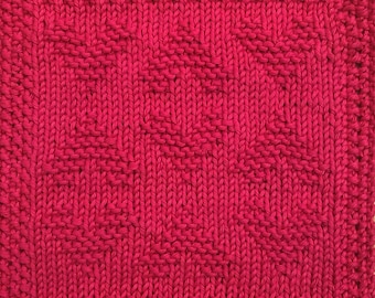 May include: A close-up of a knitted dishcloth in a bright pink color. The dishcloth has a textured pattern with a repeating diamond shape.