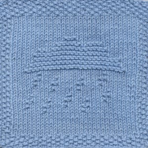Puede incluir: Una pieza cuadrada de punto azul claro con un patrón texturizado. El diseño presenta un cuadrado central con un borde en relieve y una serie de pequeñas formas en relieve dentro del cuadrado. La textura general es densa y las puntadas son claramente visibles.